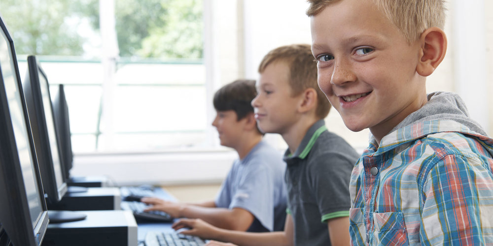 tre drenge der sidder foran hver sin computer, hvor forreste lyshårede dreng smiler til kameraet.