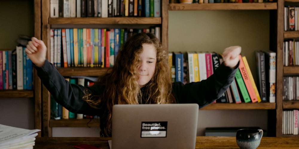 Mørkhåret pige der jubler foran sin computer med bogreol i baggrunden.